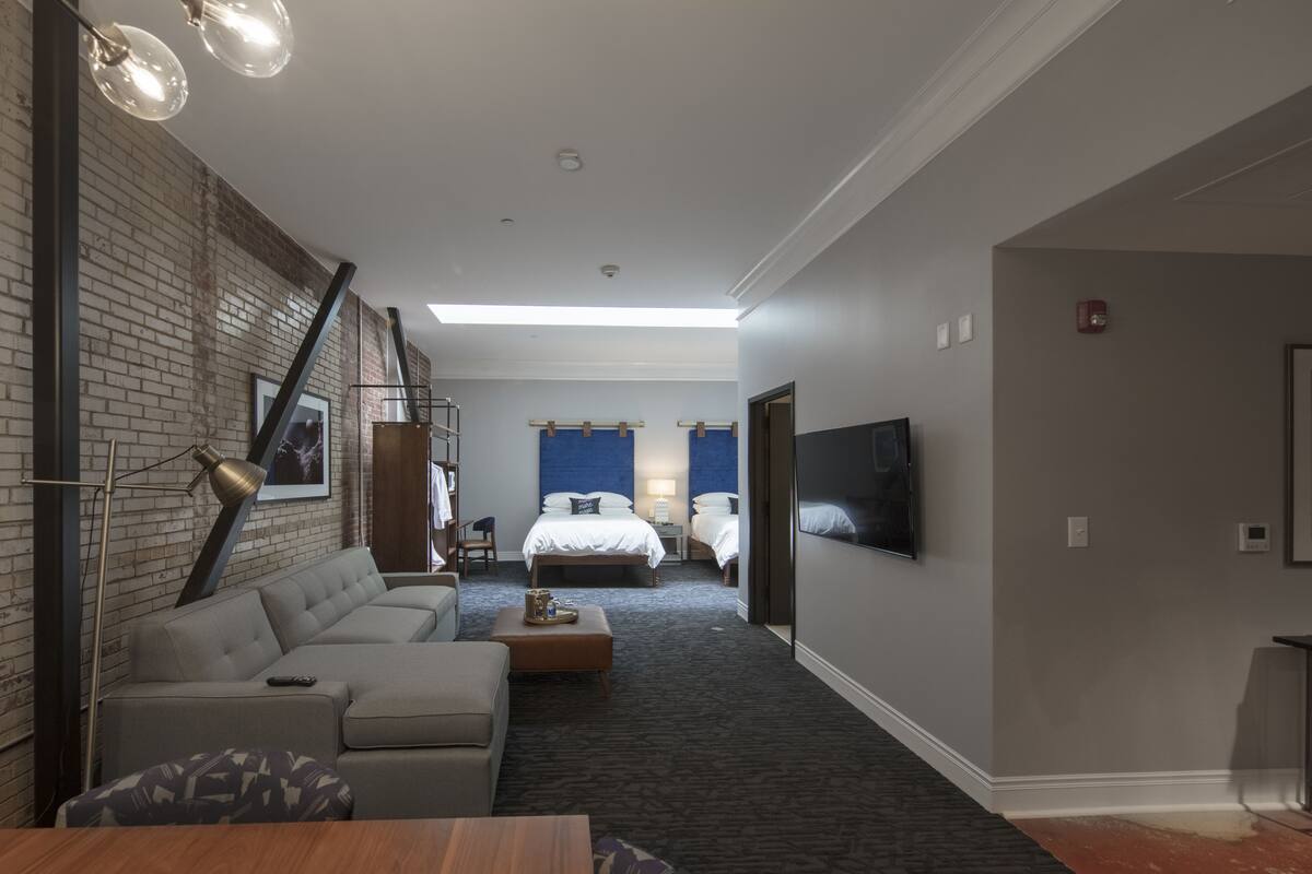 A modern hotel room with a brick accent wall, a large bed, a sofa, a desk, and a flat-screen TV.
