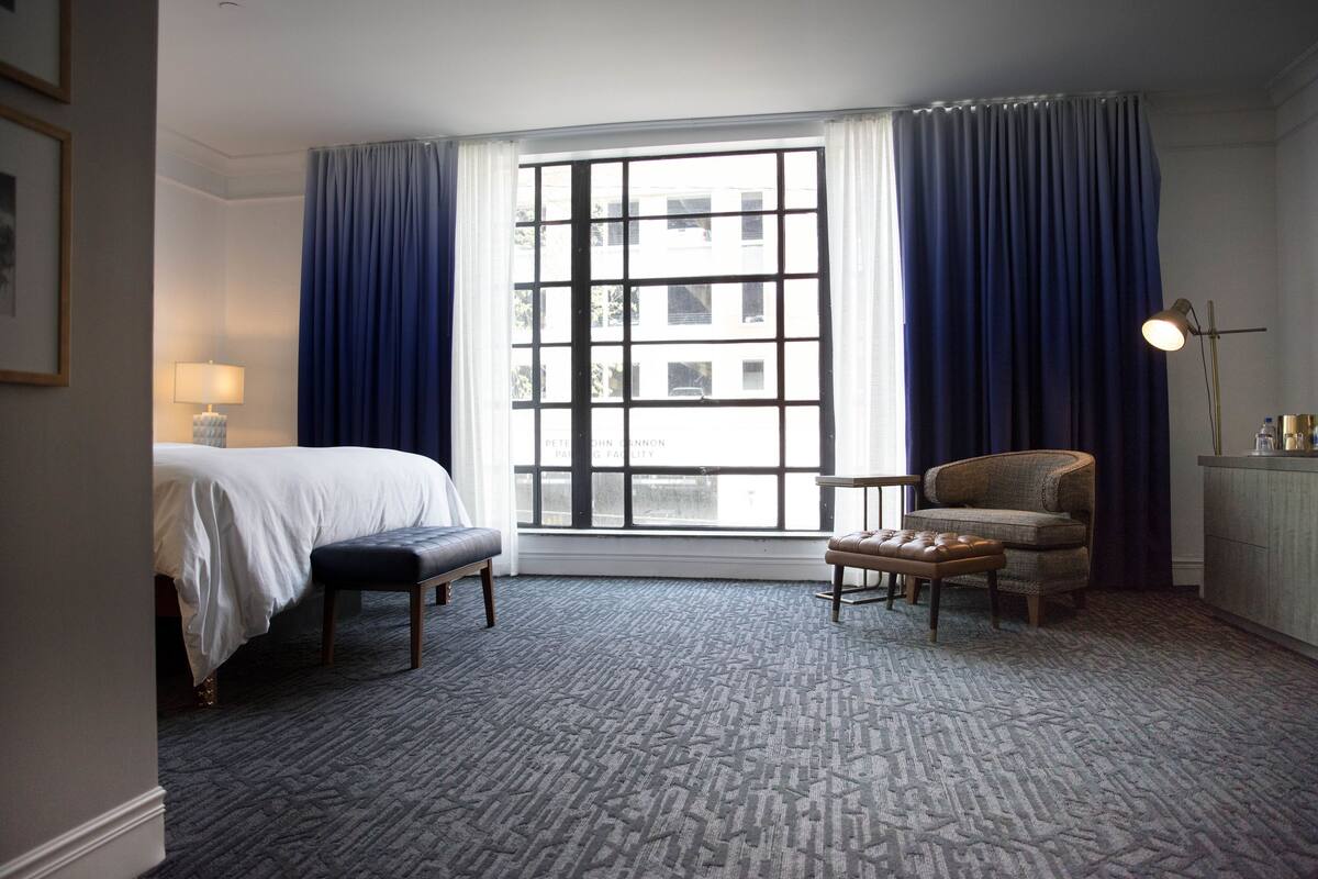 A hotel room with a bed, a chair, a small table, and a window with curtains.