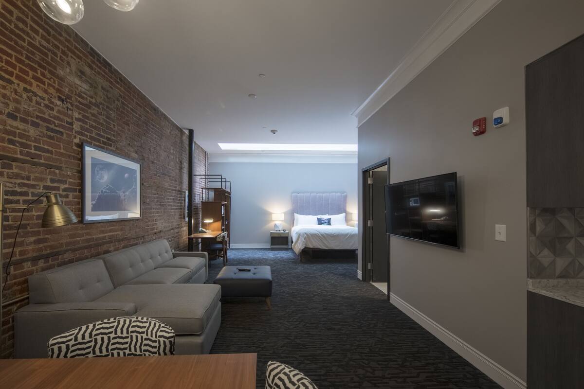 A hotel room with a brick wall, a sofa, a flat-screen TV, a bed, and a desk.