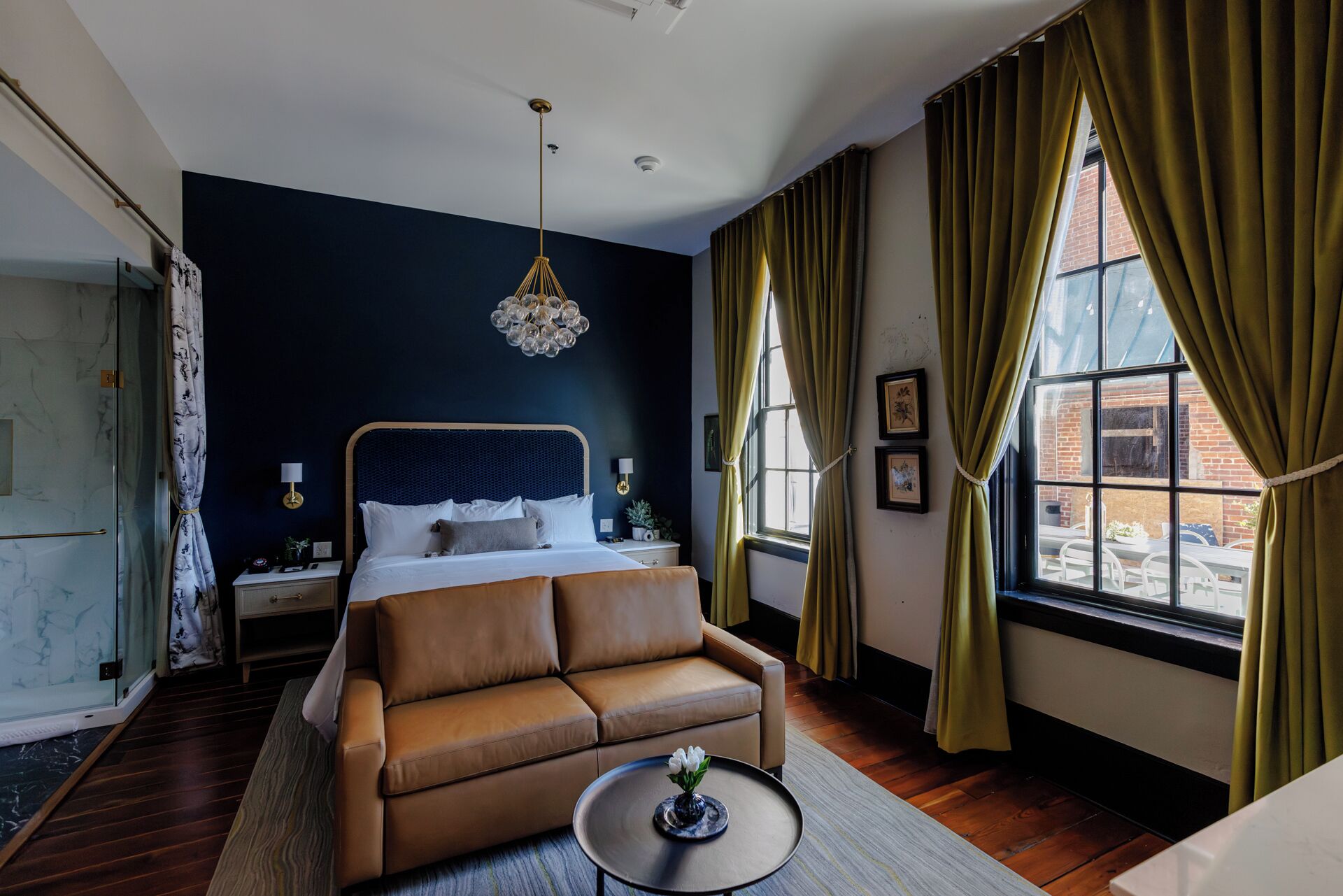Una habitación de hotel con cama, sofá, mesita de noche, lámpara, ventana con cortinas y candelabro.