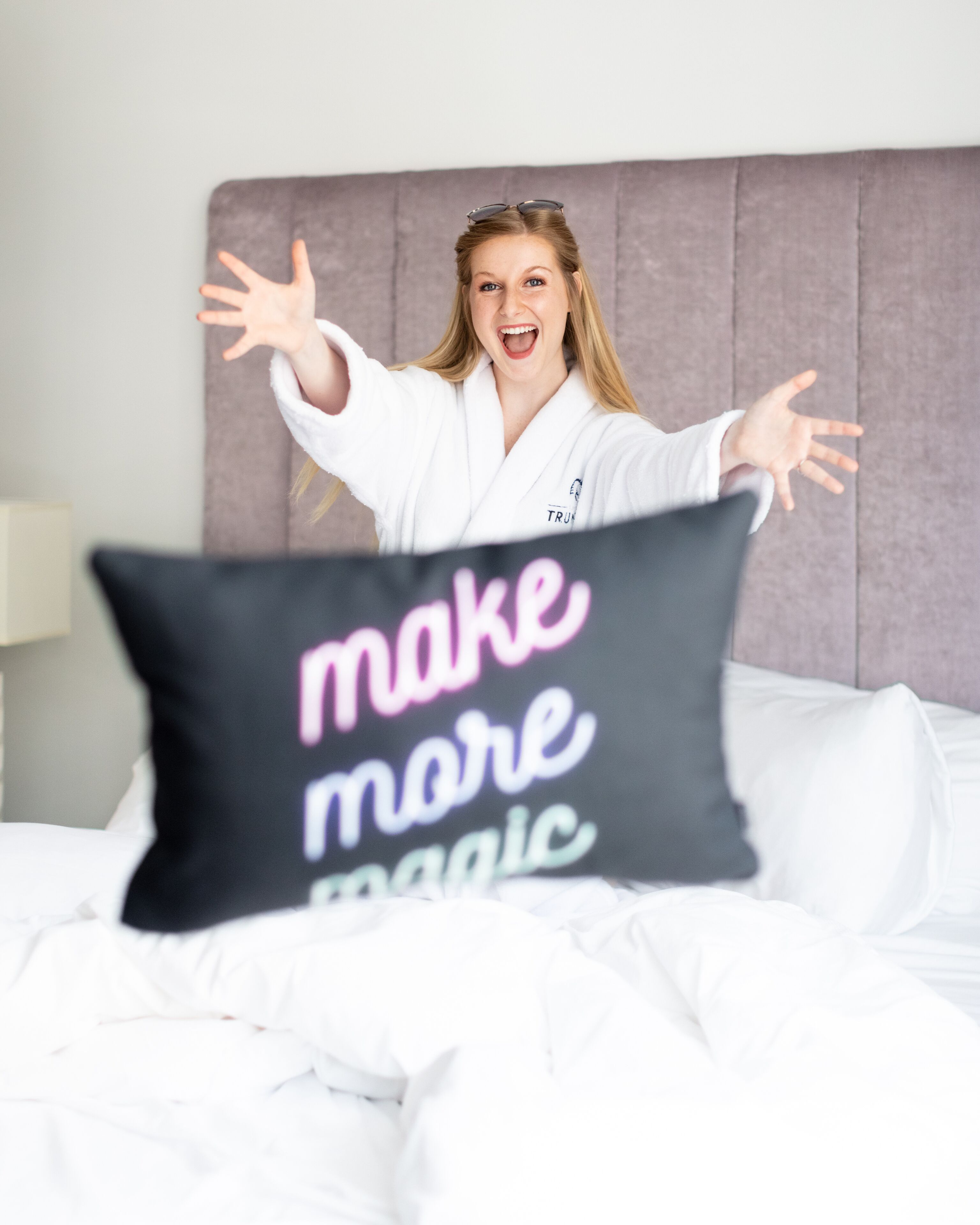 A person in a white robe sitting on a bed with a pillow that reads "make more magic."