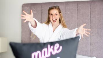 A person in a white robe sitting on a bed with a pillow that reads "make more magic."