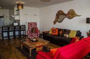 A living room with a red sofa, a black leather couch, a wooden coffee table, and a patterned armchair. There is a kitchen area with white cabinets and a dining table with chairs. - Greenways Golf Estate Sea View Apartment (Cape Town)