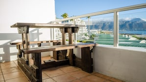 Ein Balkon mit einem rustikalen Holztisch, einer weißen Wand und Blick auf eine Küstenlandschaft mit Bergen und grünen Feldern.