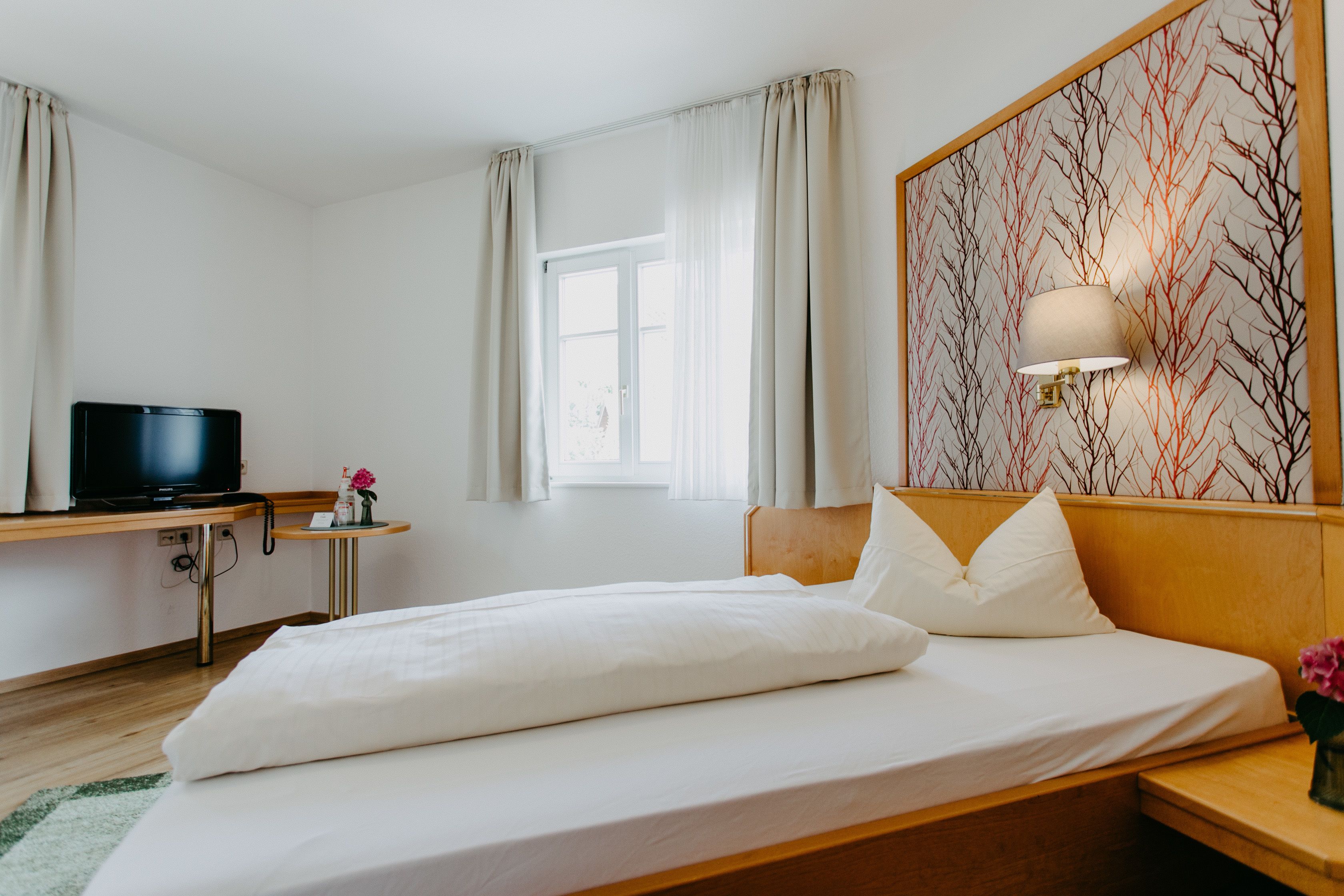 A hotel room with a bed, a TV on a desk, a window with curtains, and a wall with a tree pattern.