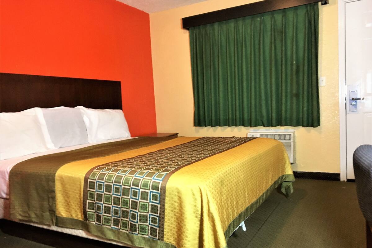 A hotel room with a bed, a window with green curtains, a radiator, and a door.