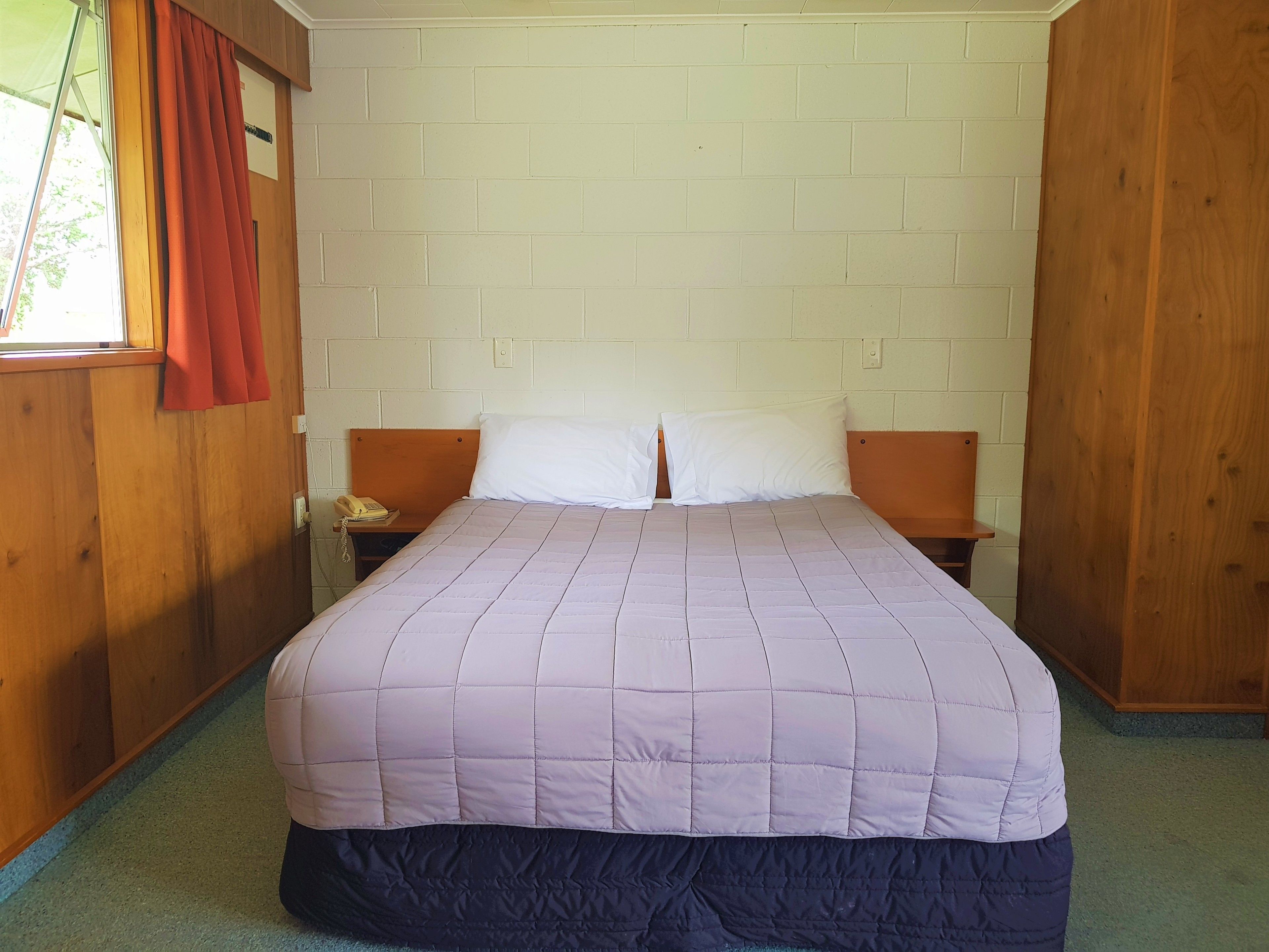 Een enkel bed met een gewatteerde spreidek, twee nachtkastjes, een houten hoofdbord en een raam met gordijn.