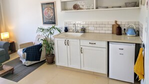A compact kitchen with white cabinets, a sink, and a dishwasher. There are shelves above the sink holding various dishes and a kettle. - Al Sfat HaBanias (HaGalil HaElyon)
