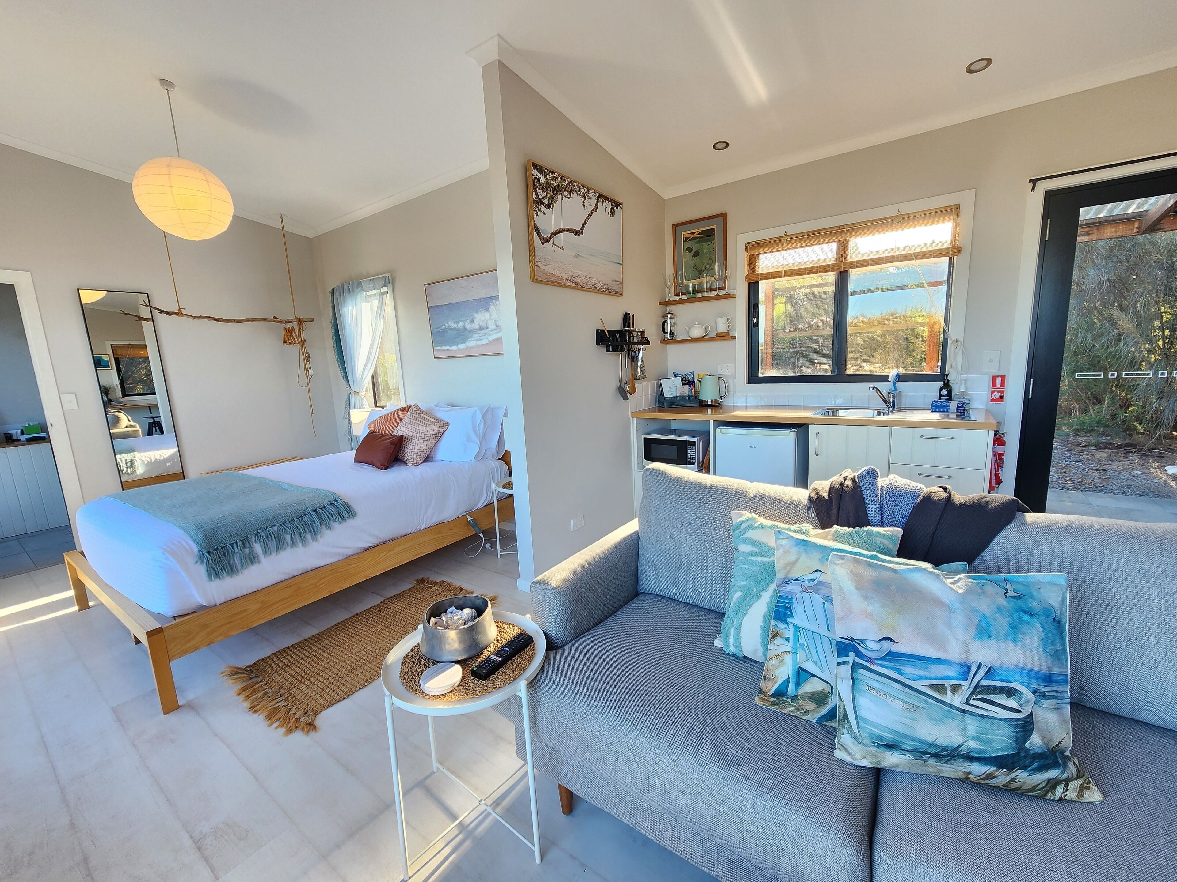 A modern bedroom with a bed, a sofa, a small table with a bowl and remote, and a kitchen area with a sink and appliances.