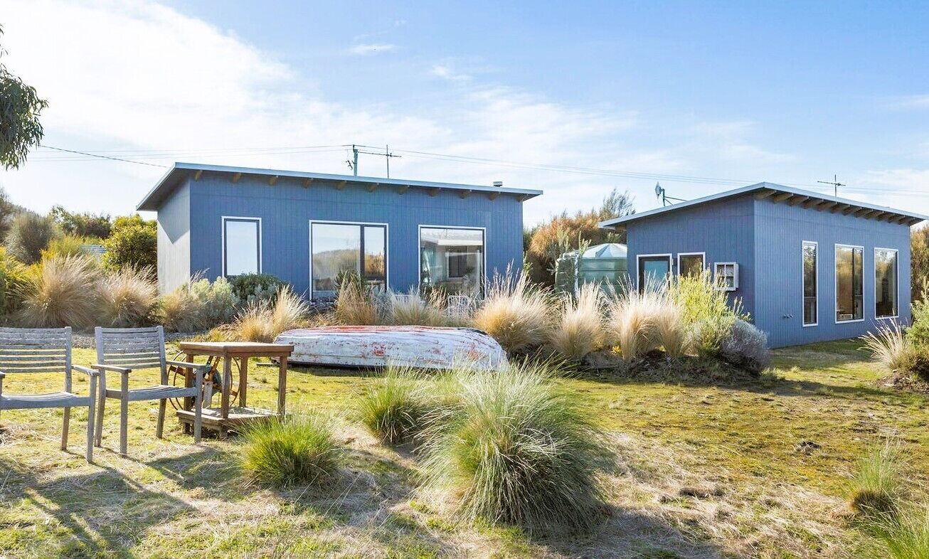 A modern two-story house with a blue exterior, large windows, and a grassy yard with outdoor furniture.