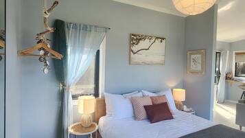 A bedroom with a bed, two bedside tables, a hanging light fixture, a window with sheer curtains, and wall art.