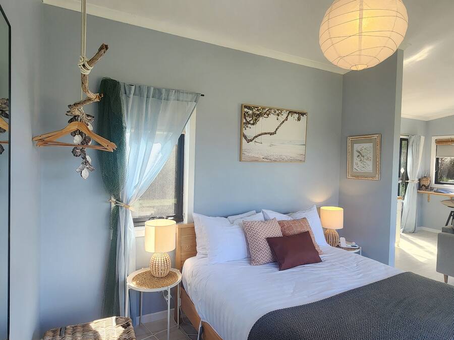 A bedroom with a bed, two bedside tables, a hanging light fixture, a window with sheer curtains, and wall art.