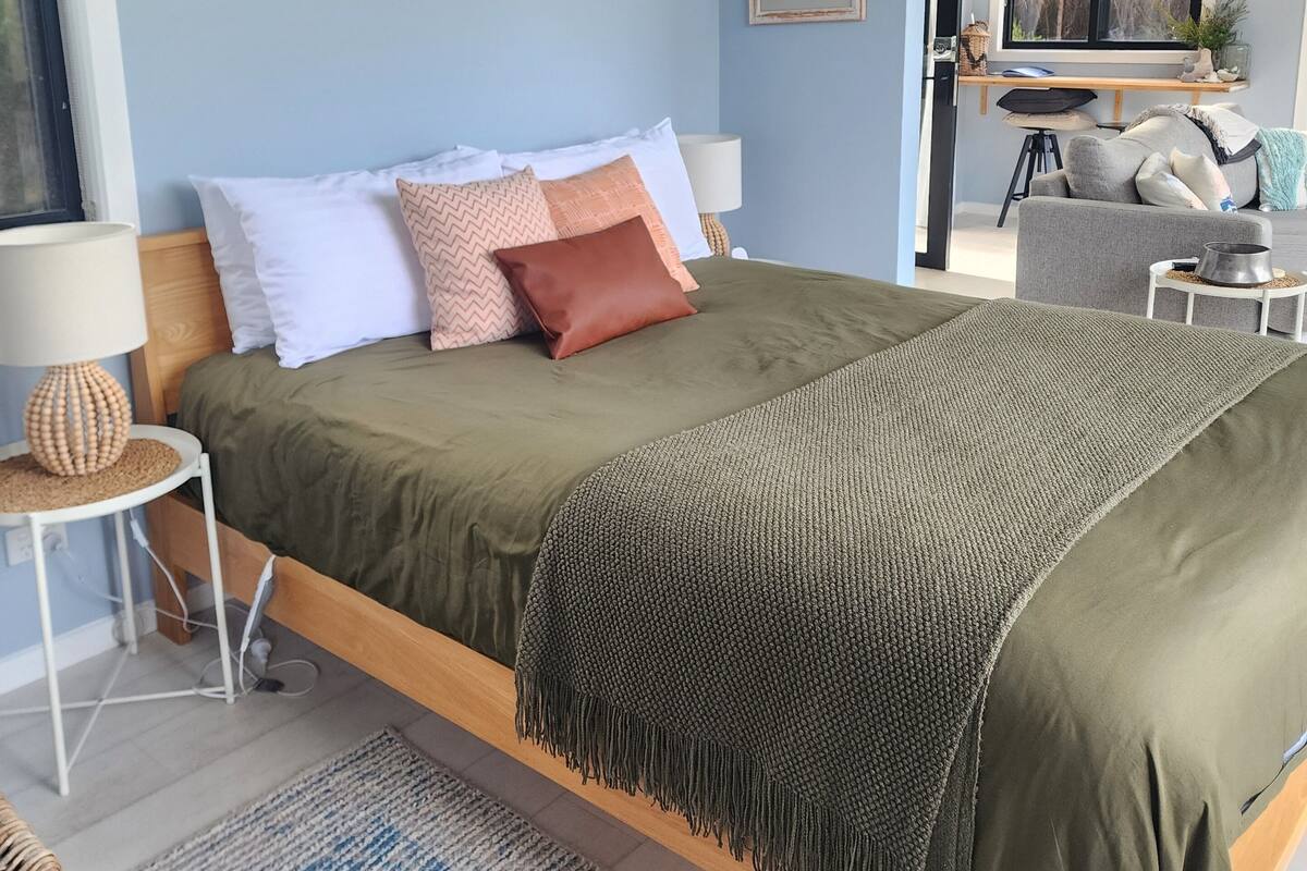 A modern bedroom with a large bed, a bedside table, a lamp, and a view of the living area.