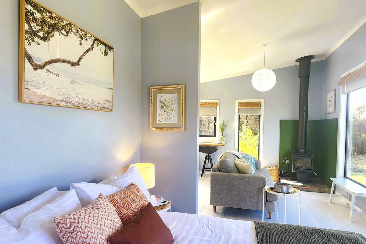 A bedroom with a bed, pillows, a bedside table, a lamp, and a view of a living area with a sofa, a table, a chair, and a fireplace.