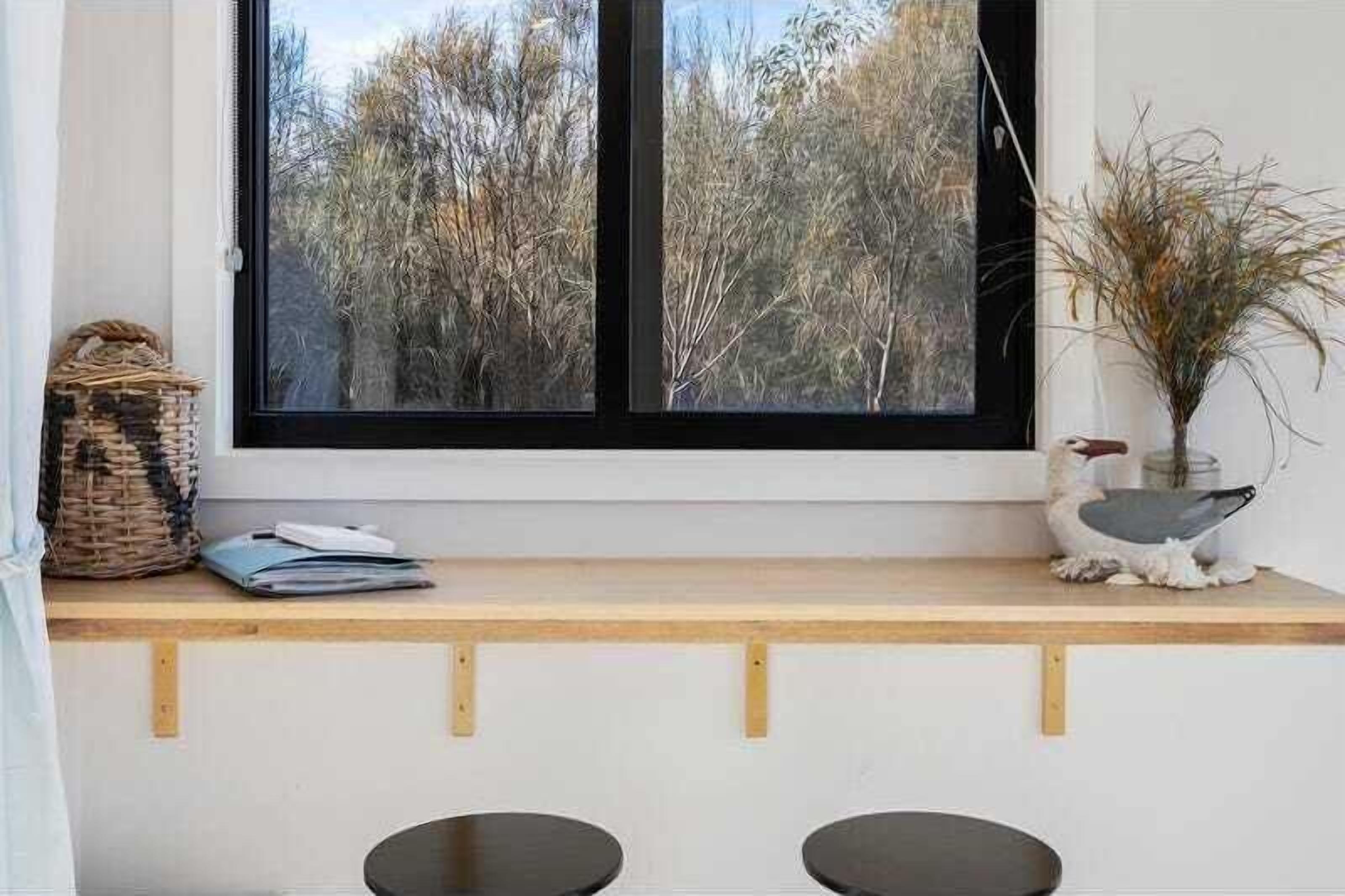 Una habitación con un amplio ventanal que da a un bosque, un banco de madera con una cesta y un florero con plantas secas, y una pequeña mesa con una libreta y un bolígrafo.