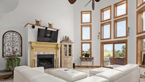 Ein geräumiges Wohnzimmer mit einem großen, weißen Ecksofa, einem Kamin mit Sims, einem Fernseher und einem großen Fenster mit vielen Scheiben.