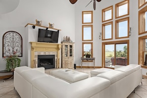A spacious living room with a large white sectional sofa, a fireplace with a mantel, a television, and a large window with multiple panes.