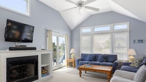 A living room with a fireplace, a television, a blue sofa, and a ceiling fan. - Lion On The Sand: Oceanside 6-bedroom, Private Pool & Hot Tub (Corolla)