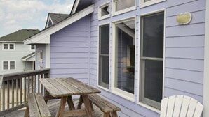 A wooden deck with a picnic table and a white chair, adjacent to a house with purple siding and white trim. - Lion On The Sand: Oceanside 6-bedroom, Private Pool & Hot Tub (Corolla)