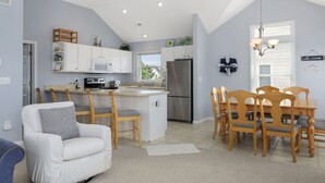 A modern kitchen with white cabinetry, a central island, and a dining area with wooden chairs and a table. - Lion On The Sand: Oceanside 6-bedroom, Private Pool & Hot Tub (Corolla)