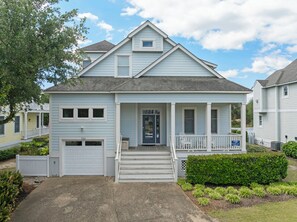 A light blue two-story house with a front porch, a garage, and a small front yard with a hedge and plants.