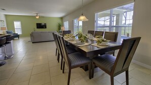 Un comedor con una mesa larga dispuesta para seis personas, rodeada de sillas. En el fondo, un televisor está montado en la pared, y hay un sofá y una pequeña mesa con sillas en la zona de estar contigua.