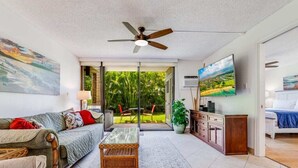 Uma sala de estar espaçosa com sofá, mesa de centro e TV. Há um ventilador de teto, uma janela grande com vista para o verde e uma porta deslizante que dá para uma área externa.