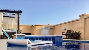 Un área de piscina en la azotea con una piscina azul cristalina, un sillón flotante blanco y una pérgola con cortinas blancas.