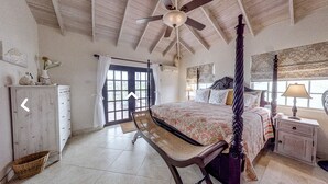 Un dormitorio con una cama con dosel, un ventilador de techo, una ventana con cortinas, una mesita de noche con una lámpara y una silla de mimbre.