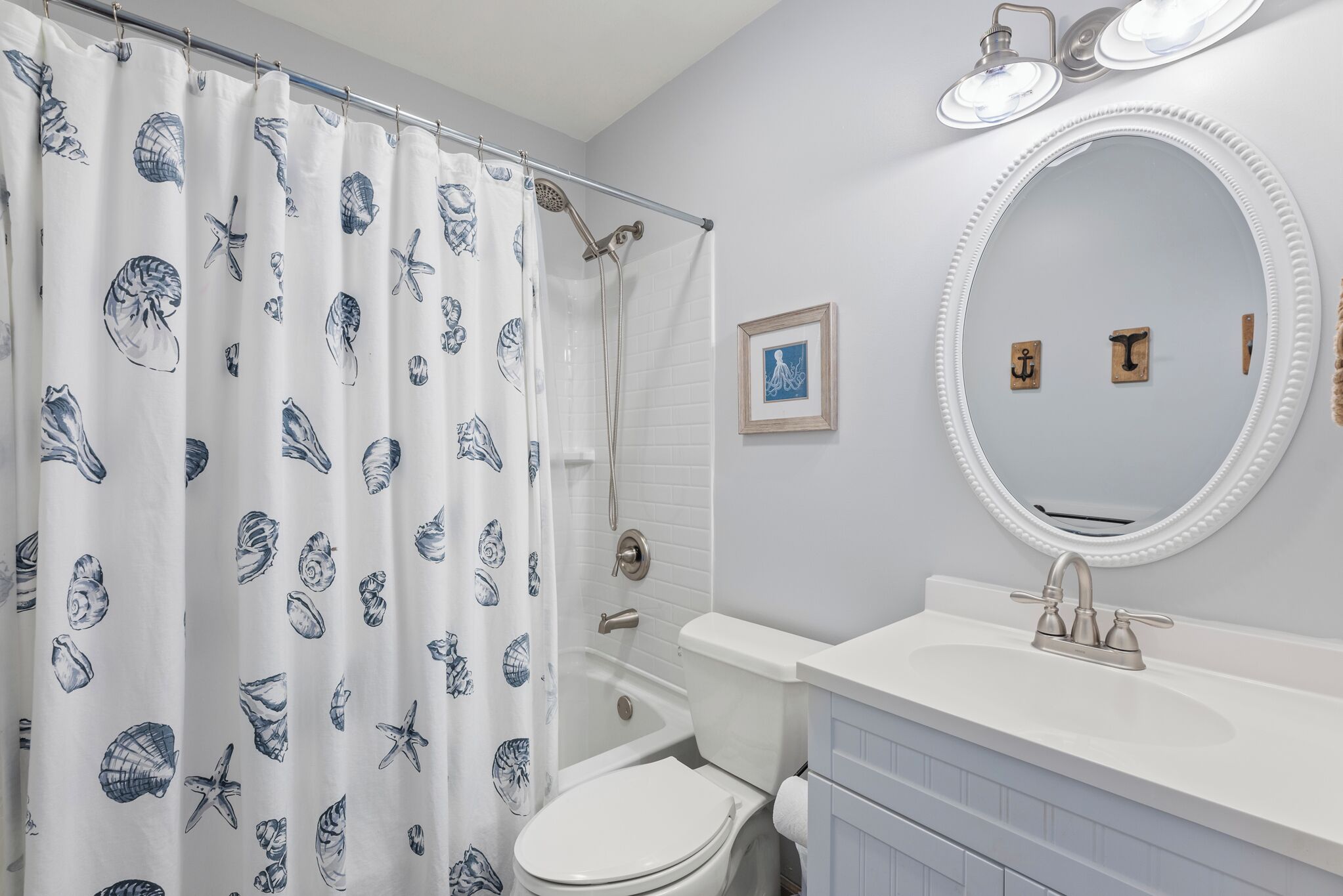 Ein Badezimmer mit Toilette, einer Badewanne, einem Duschvorhang mit Muschelmuster, einem weißen Waschbecken mit Spiegel darüber und einem gerahmten Bild an der Wand.