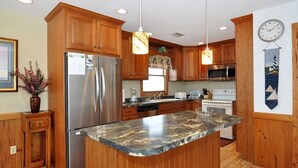 A kitchen with wooden cabinets, stainless steel appliances, a marble countertop, pendant lighting, and a clock on the wall. - Merry Winds: Oceanfront, Shared Pool, Great Views (Corolla)