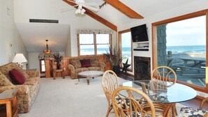 A spacious living room with a large window overlooking the ocean, a fireplace, and a dining area with chairs and a table. - Merry Winds: Oceanfront, Shared Pool, Great Views (Corolla)