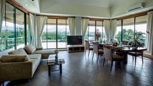 A spacious living room with a sofa, dining table, and chairs, large windows with curtains, a flat-screen TV, and a balcony with a view. - Khaoyai Fahsai Resort (Pak Chong)