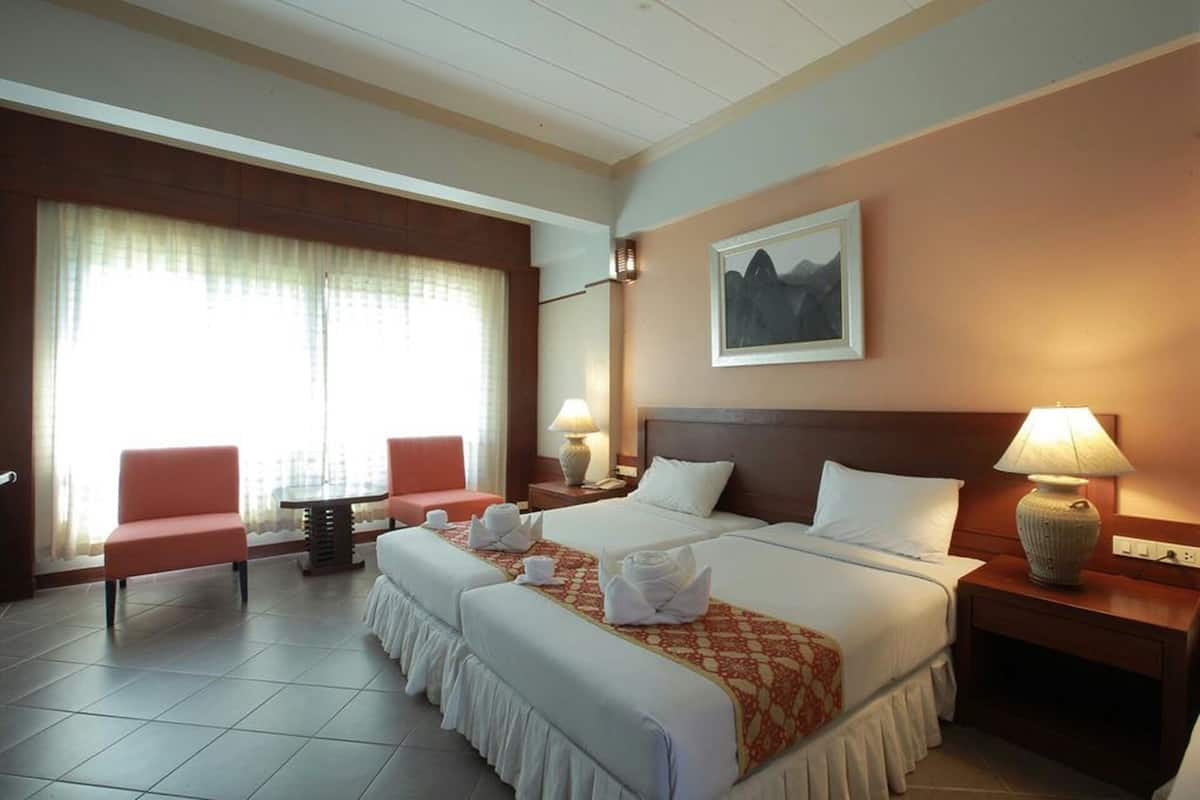 Ein Hotelzimmer mit zwei Betten, einer Sitzecke inklusive Sessel und Tisch sowie einem großen Fenster mit Vorhängen.