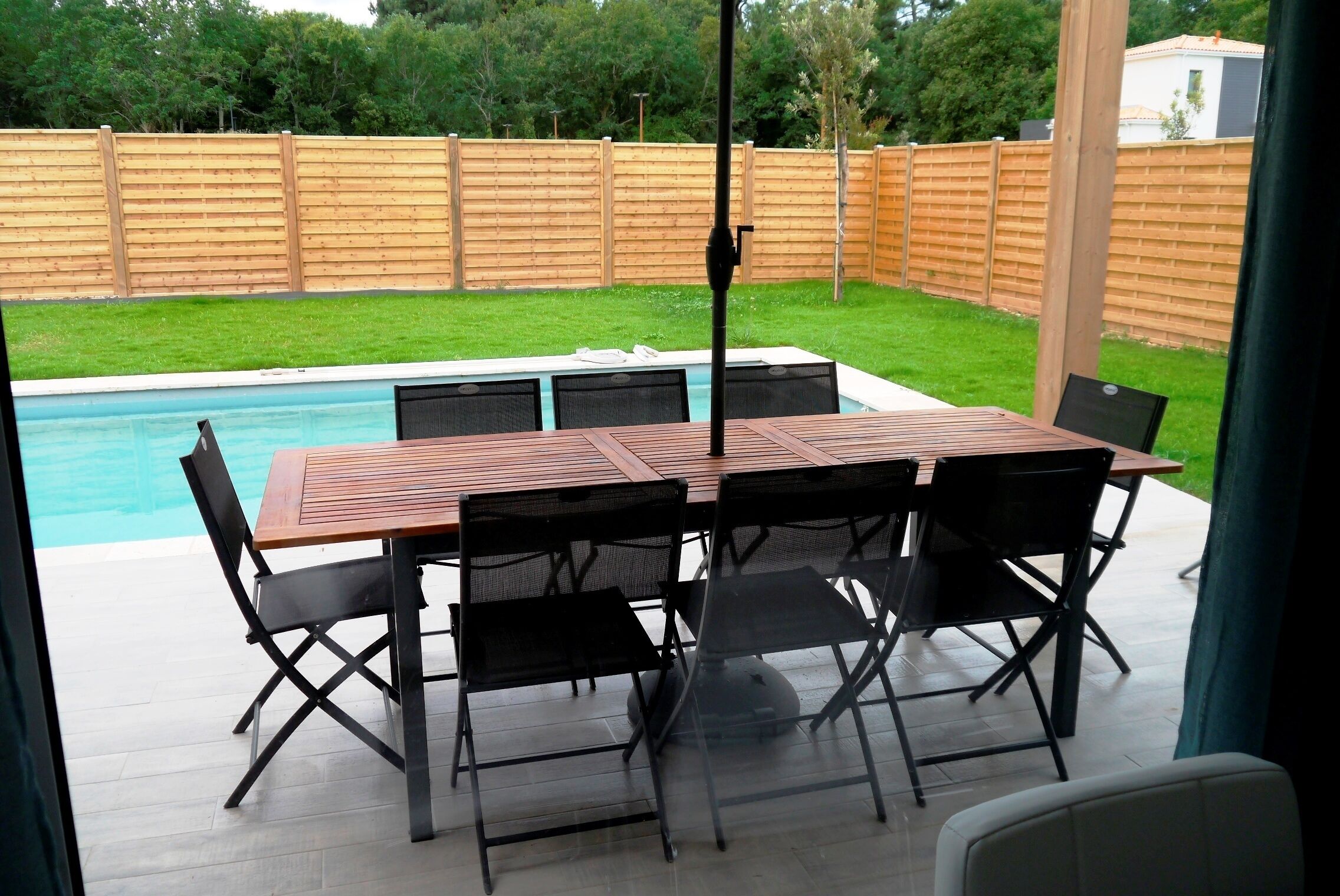 Une terrasse extérieure avec une table en bois et des chaises noires, une piscine et une clôture en bois en arrière-plan.