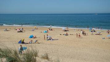 Une plage de sable où l’on peut voir des gens se prélasser au soleil et se baigner, des parasols et un ciel bleu azur.