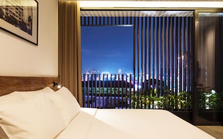 A modern hotel room with a large bed, a city view through floor-to-ceiling windows, and a framed picture on the wall.
