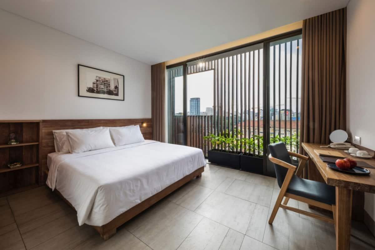 A modern hotel room with a large bed, a desk with a chair, a view through large windows, and a framed picture on the wall.