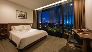 A hotel room with a large bed, a desk with a chair, a view of the city at night, and a balcony with plants. - Triple E Hotel Fine Art Museum (Ho Chi Minh City)