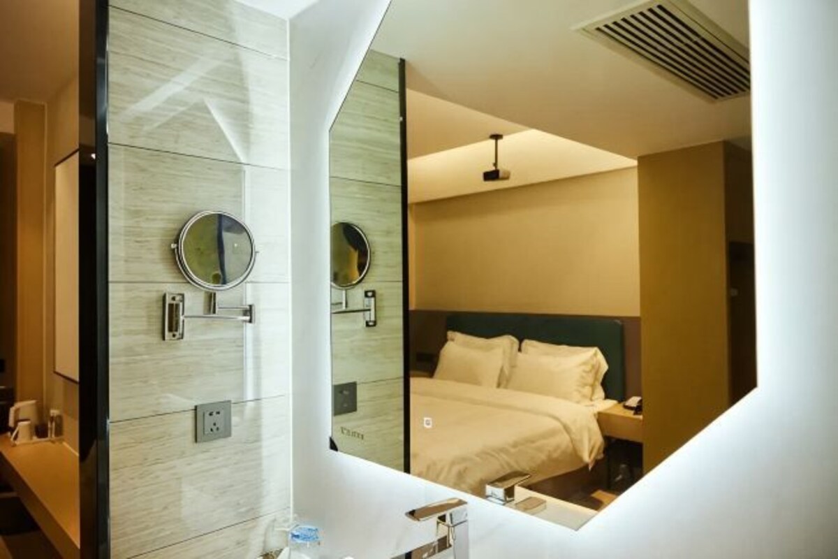 A modern hotel room with a large mirror, a bed with white linens, a bedside table, and a wall-mounted light fixture.