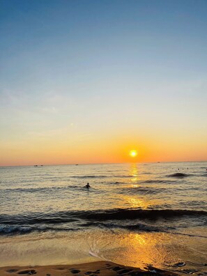 Une scène de plage avec le soleil se couchant sur l’océan, une eau calme et une rive sablonneuse.