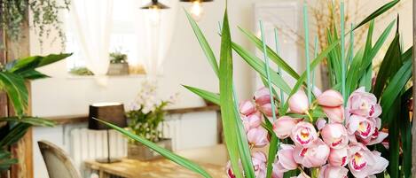 A cozy dining area with a wooden table and chairs, surrounded by potted plants and a vase of pink flowers.