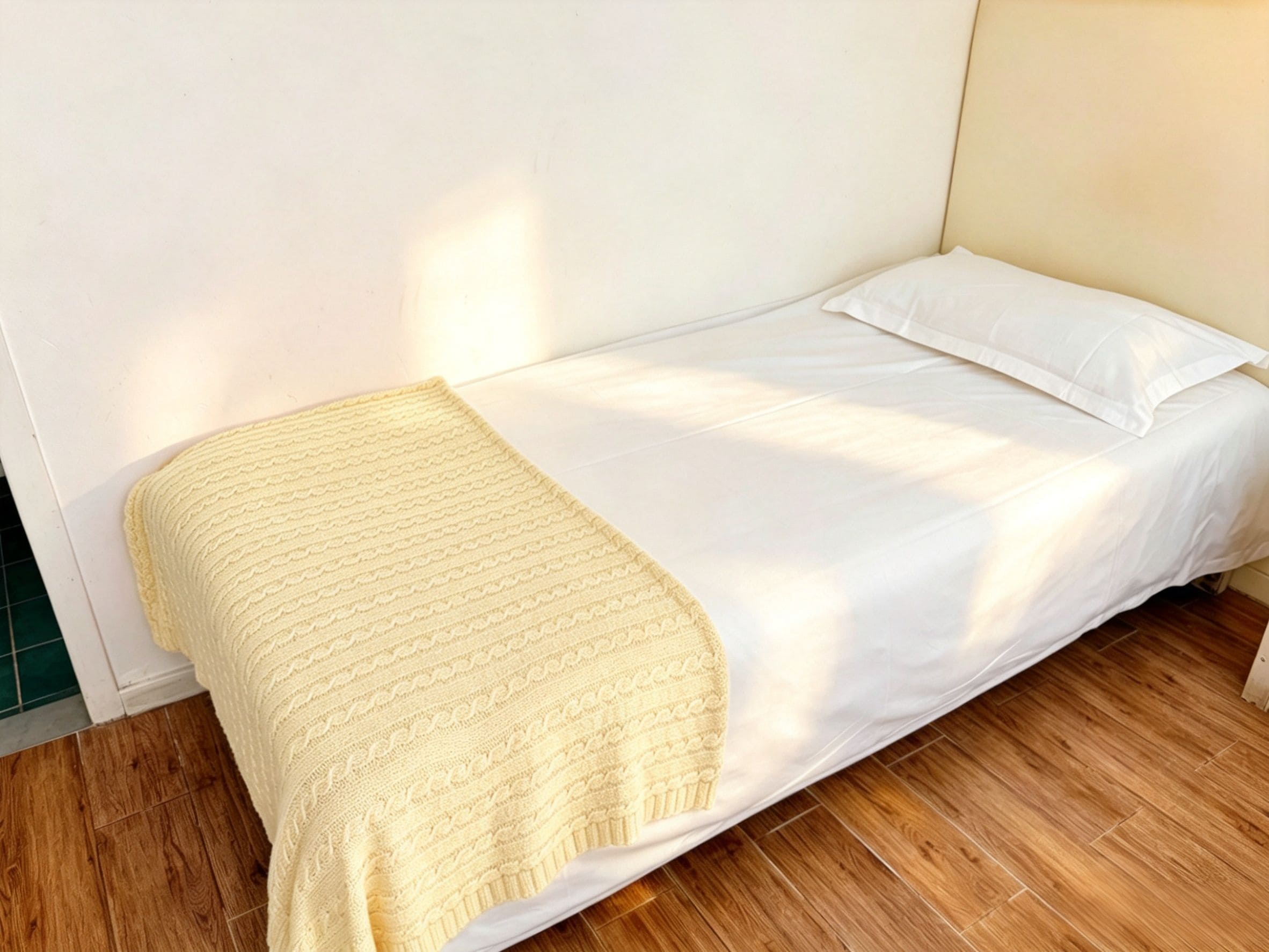 A single bed with a white mattress and a yellow blanket in a room with wooden flooring and white walls.