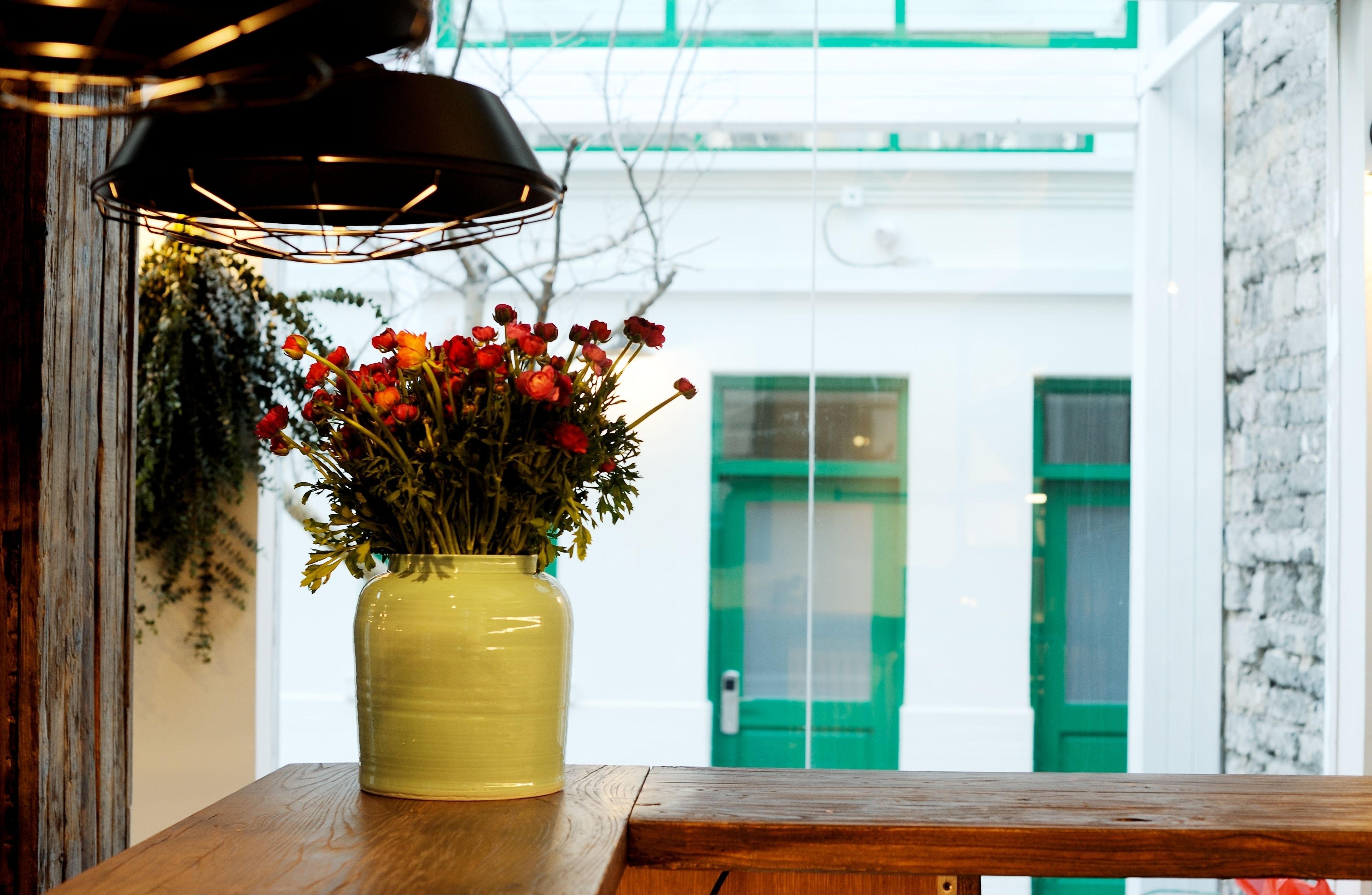 A yellow vase with red flowers on a wooden table, a green door, and a white wall with a window in the background.