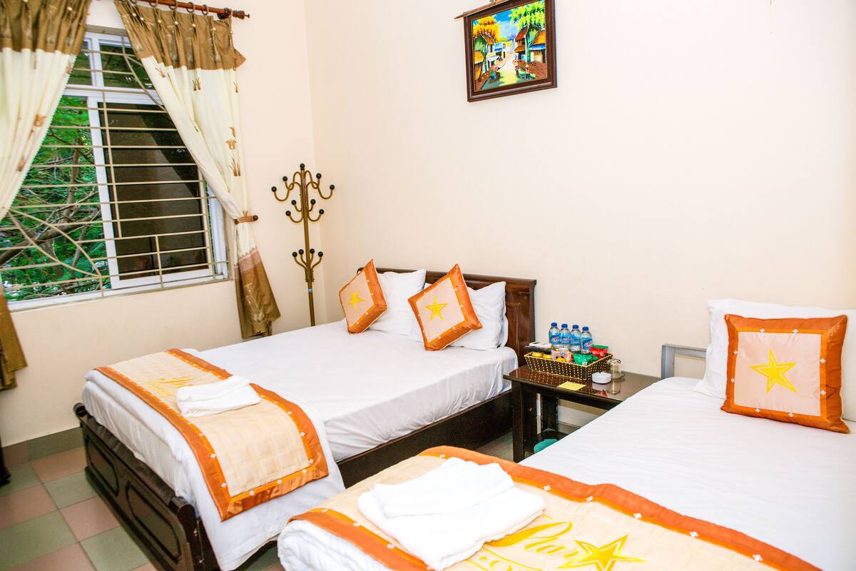 A hotel room with two beds, a window with curtains, a painting on the wall, and a small table with toiletries.