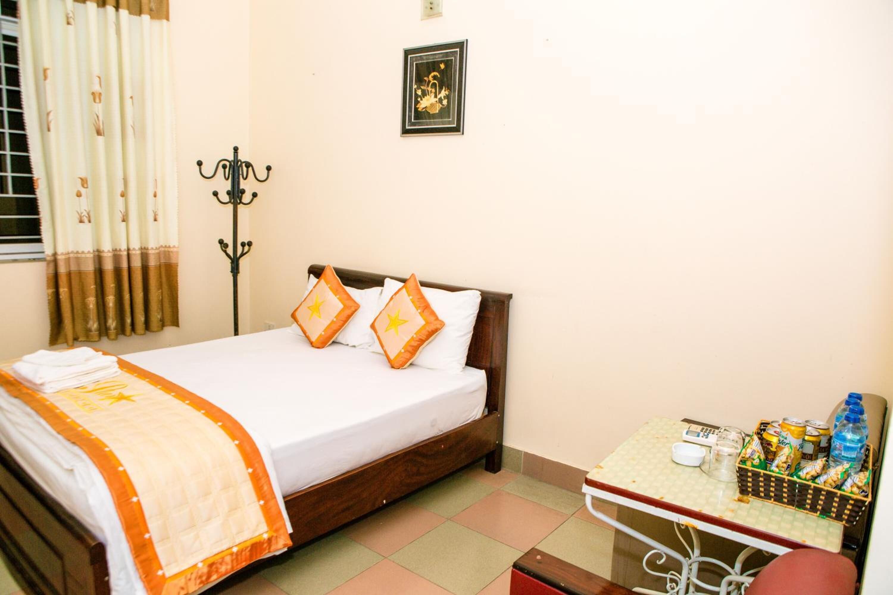 A neatly made bed with a white and orange bedspread, two pillows with star designs, a small table with snacks and drinks, and a wall-mounted coat rack.