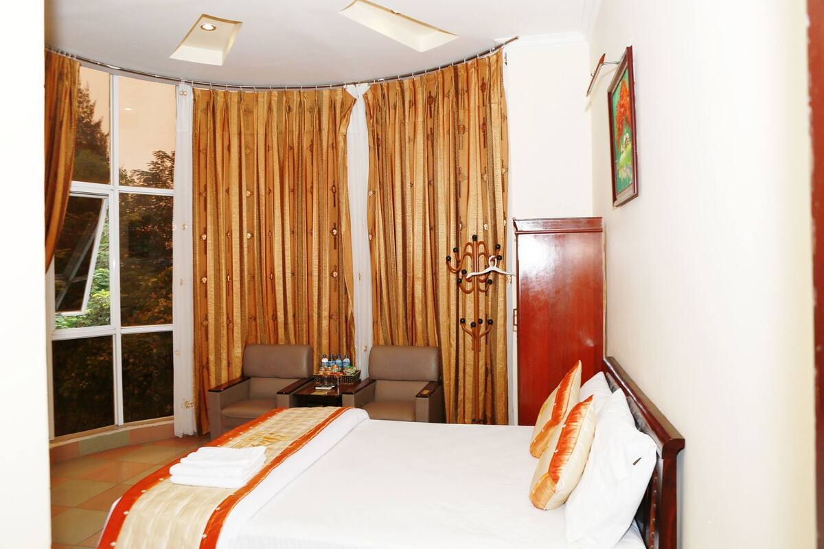 A hotel room with a bed, two armchairs, a small table with bottles, a wooden cabinet, and a large window with curtains.
