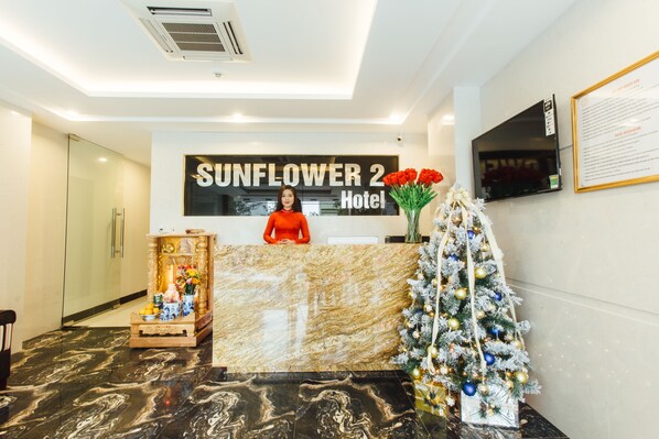 Bancada de recepção do Hotel Sunflower 2, com bancada de mármore, uma árvore de Natal e um cardápio emoldurado na parede.