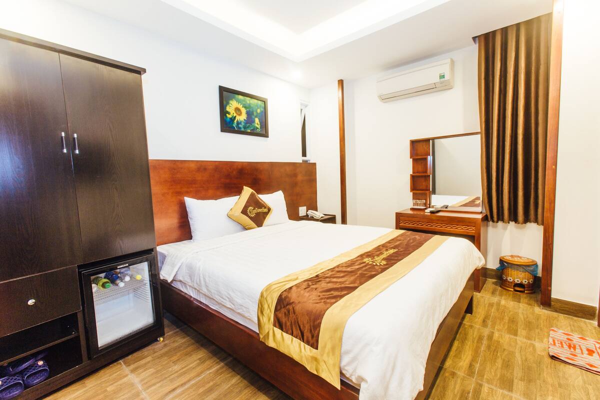 Habitación de hotel con un armario grande, una cama con ropa de cama blanca y detalles marrones, un refrigerador pequeño, una mesita de noche con lámpara y un cuadro en la pared.
