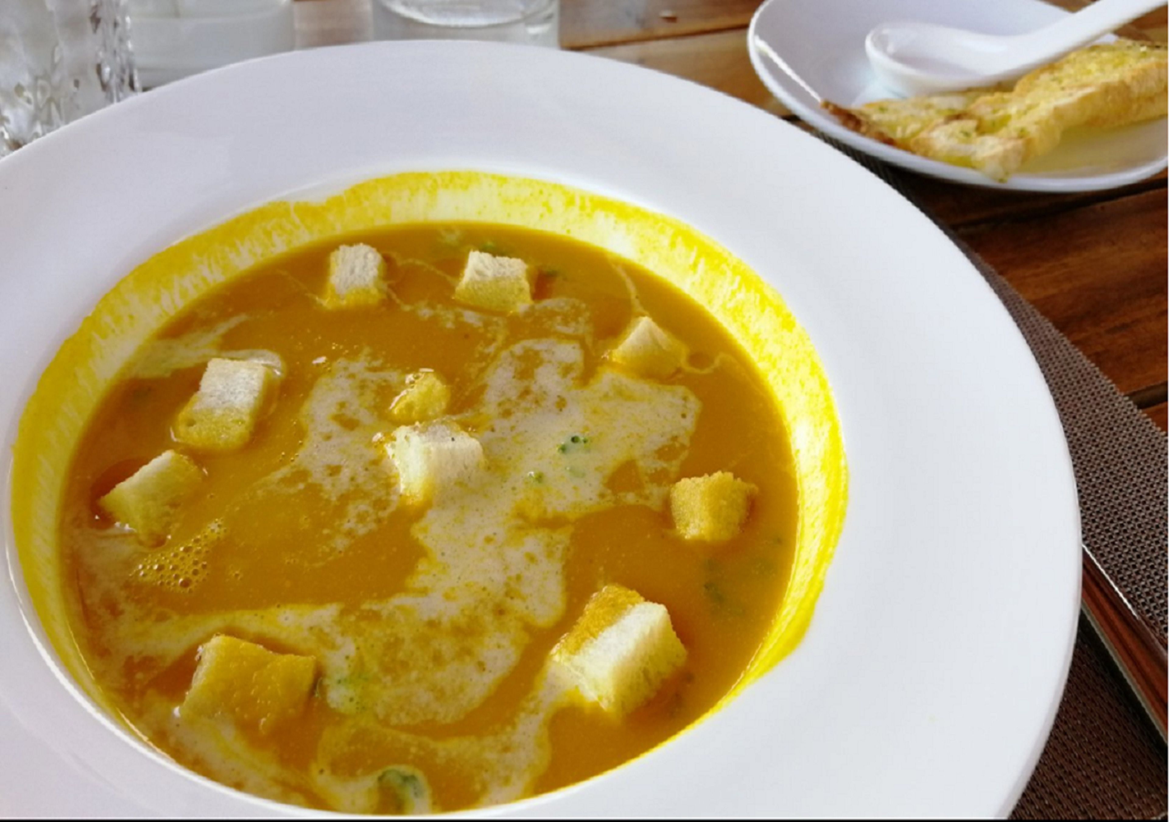 A bowl of soup with cubes and a spoon on a plate.